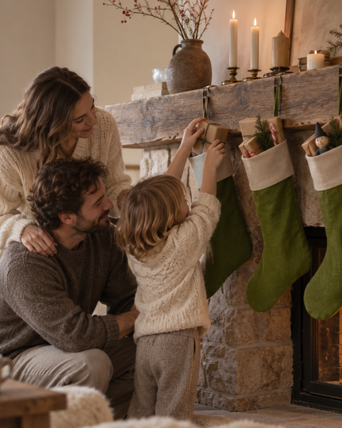 A cozy family scene by a fireplace with a woman, man, and child hanging green Christmas stockings under a rustic mantel with lit candles.