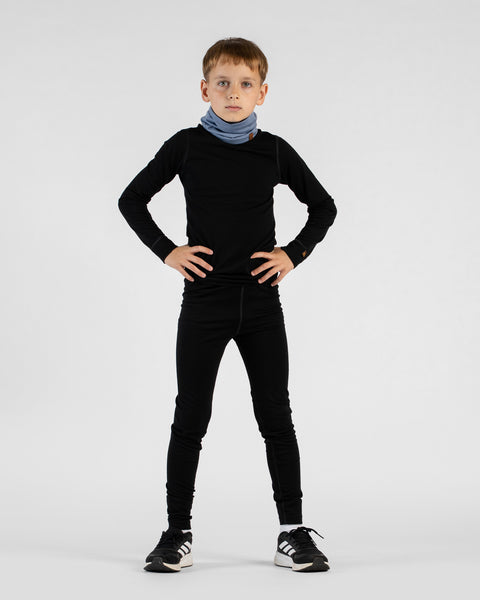 A young boy stands facing forward with his hands on his hips against a white background. He is wearing a black long-sleeved base layer top, black leggings, black and white Adidas sneakers, and a light blue neck gaiter.