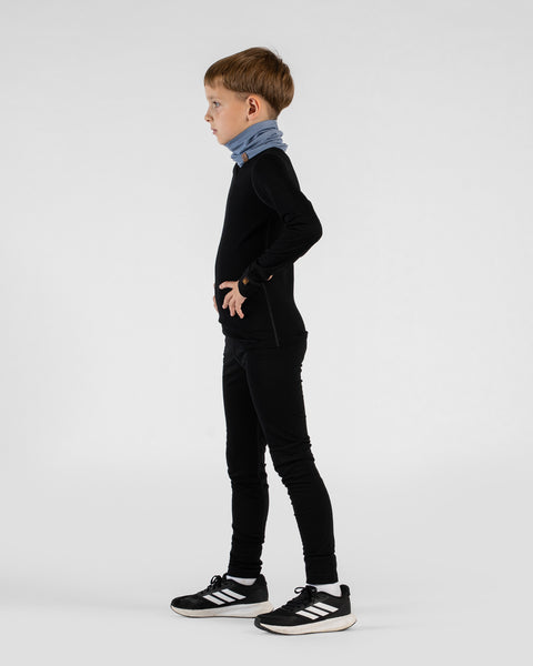 A young boy stands in profile facing left with his hands on his hips against a white background. He is wearing a black long-sleeved base layer top, black leggings, black and white Adidas sneakers, and a light blue neck gaiter.