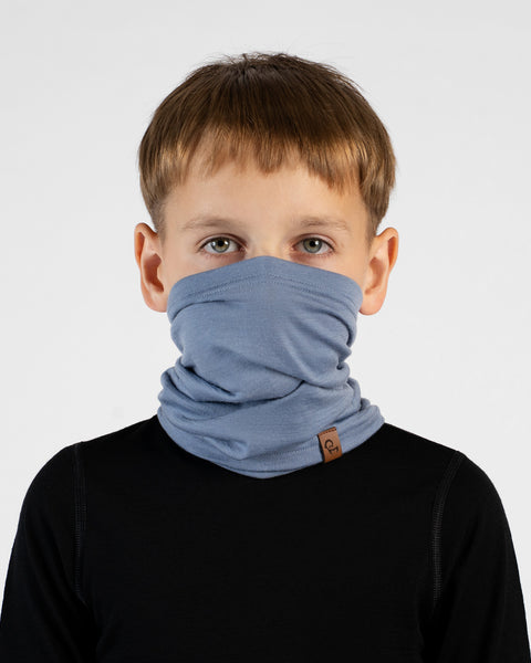 A close-up portrait of a young boy looking directly at the camera against a white background. He is wearing a black long-sleeved top and a light blue neck gaiter pulled up to cover his mouth and nose, with a small brown logo tag visible.