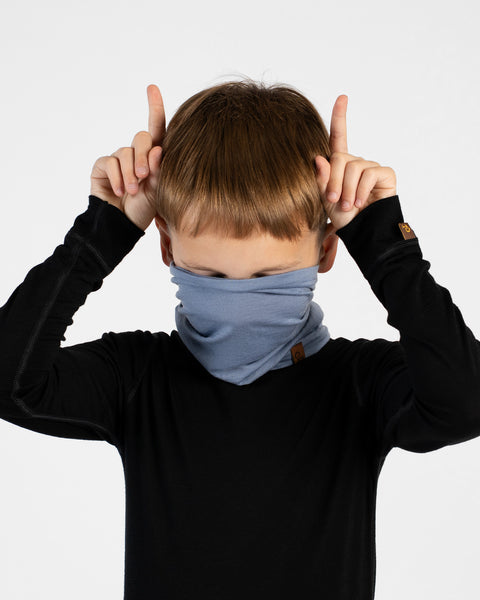 A young boy stands facing forward against a white background, making a playful gesture with his hands by raising his index fingers behind his head like horns. He is wearing a black long-sleeved top and a light blue neck gaiter pulled up to cover his mouth and nose.