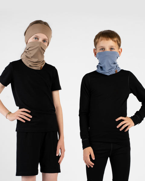 A studio portrait of two children standing side-by-side against a white background, both looking directly at the camera with one hand resting on their hip. The girl on the left is wearing a black short-sleeved t-shirt, black shorts, a beige headband, and a matching beige neck gaiter pulled up over her nose and mouth. The boy on the right is wearing a black long-sleeved base layer top, black leggings, and a light blue neck gaiter pulled up to cover his nose and mouth.