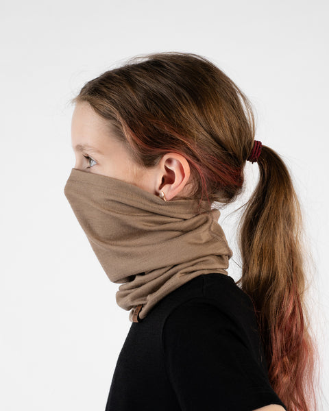 A close-up studio portrait of a young girl standing in profile facing left against a white background. She is wearing a black short-sleeved top and a beige neck gaiter pulled up to cover her mouth and nose. Her hair is pulled back in a ponytail with a red hair tie.