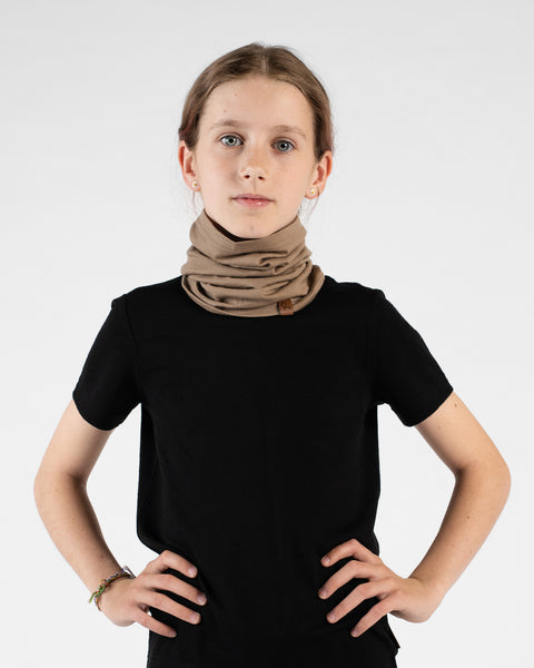 A studio portrait of a young girl standing with her hands on her hips, looking directly at the camera against a white background. She is wearing a black short-sleeved t-shirt and a beige neck gaiter bunched loosely around her neck, featuring a small brown logo tag.