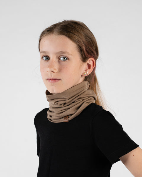 A close-up portrait of a young girl looking directly at the camera in a three-quarter profile against a white background. She is wearing a black short-sleeved top and a beige neck gaiter bunched around her neck, featuring a small brown logo tag.
