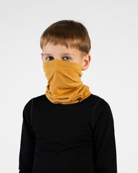 A close-up portrait of a young boy looking directly at the camera against a white background. He is wearing a black long-sleeved top and a mustard yellow neck gaiter pulled up to cover his mouth and nose, with a small brown logo tag visible.