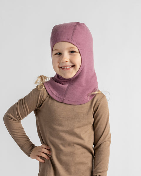 A young child wearing a dusty rose knit full-head hood or balaclava over a brown long-sleeve shirt, standing against a light gray studio background.