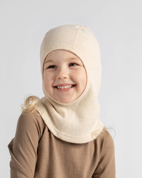 A smiling young child wearing a cream-colored knit full-head hood or balaclava over a brown long-sleeve top, photographed against a light gray studio background.