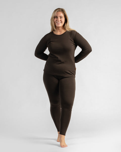 A full-body studio shot of a woman standing with hands on her hips, wearing a matching dark brown thermal long-sleeve top and leggings.