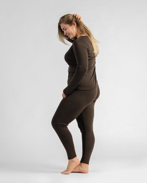 A side-profile view of a woman standing barefoot against a white backdrop, looking down while wearing fitted dark brown thermal loungewear.