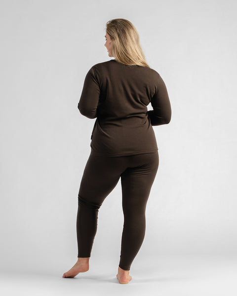 A full-length back view of a woman standing barefoot in a fitted dark brown two-piece thermal base layer set.