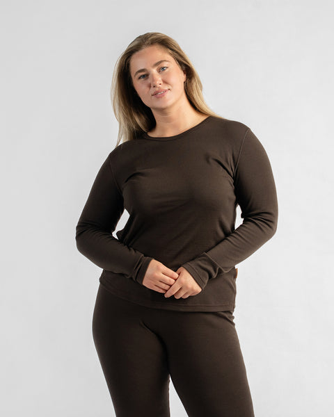 A woman standing against a neutral studio backdrop, wearing a dark brown long-sleeve thermal top and matching pants while clasping her hands at her waist.