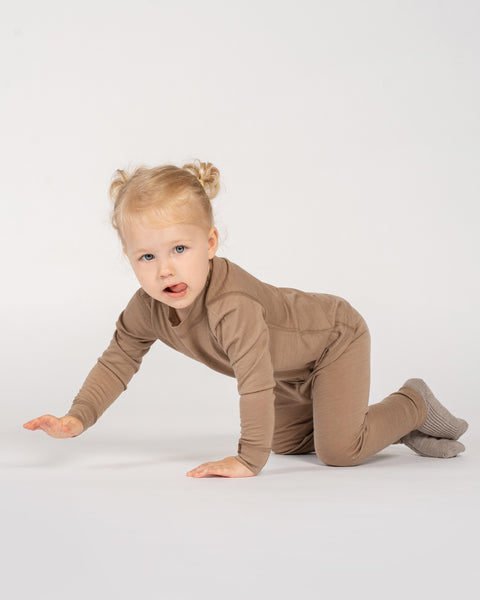 A young toddler with blonde pigtails crawls toward the camera against a plain white background, playfully sticking her tongue out. She is dressed comfortably in a matching taupe long-sleeve thermal top, leggings, and grey knit socks.