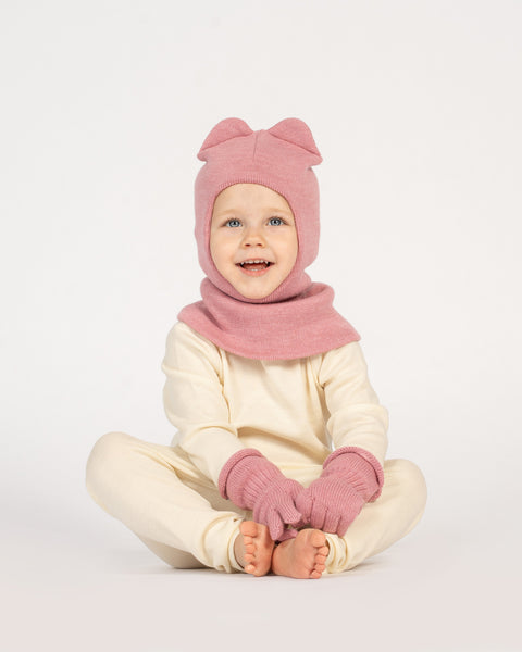 A young child with blue eyes and a wide, happy smile, is sitting cross-legged on a white surface. They are wearing a pink balaclava with ears, matching pink mittens, and a light-colored long-sleeved shirt and pants.