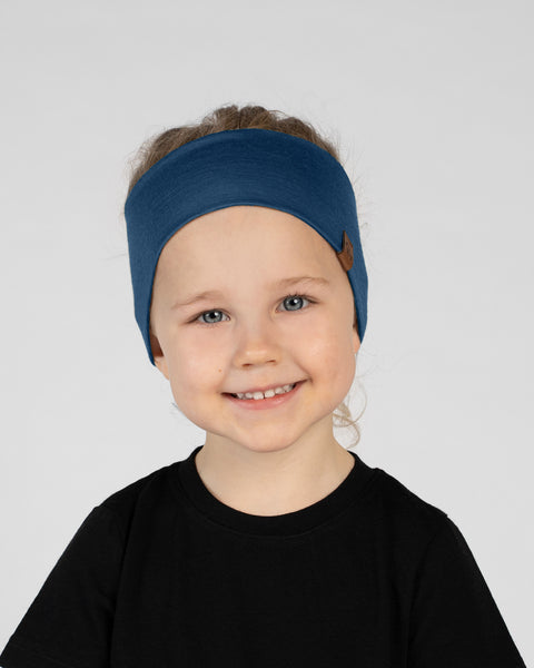 A close-up studio portrait of a smiling young child looking directly at the camera against a plain light grey background. She is wearing a black short-sleeved t-shirt and a wide denim blue headband with a small brown logo tag on the side.