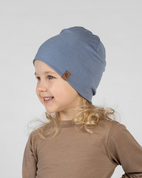 A studio portrait of a smiling young blonde girl looking off to the left side of the frame. She is wearing a close-fitting dusty blue beanie hat featuring a small brown leather logo patch on the front edge. Her blonde curls rest on the shoulders of her light brown long-sleeved shirt. The background is a plain light grey.