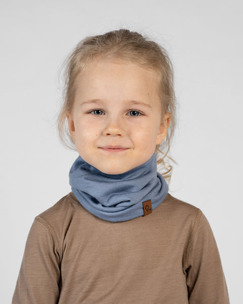 A studio portrait of a young blonde girl looking directly at the camera with a subtle, sweet smile. Her hair is loosely pulled back, and she is wearing a dusty blue neck gaiter gathered around her neck. The gaiter features a small brown leather logo patch near the bottom edge. She is wearing a light brown long-sleeved shirt against a plain light grey background.