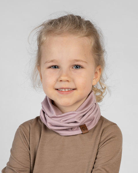 A studio portrait of a young blonde girl looking directly at the camera with a happy smile. Her hair is pulled back, and she is wearing a dusty pink neck gaiter gathered comfortably around her neck. The gaiter has a small brown leather logo patch on the lower edge. She is wearing a light brown long-sleeved shirt against a plain light grey background.