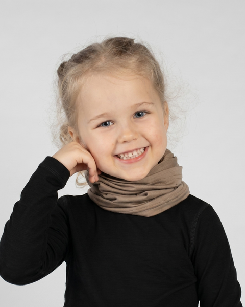 A studio portrait of a young blonde girl looking directly at the camera with a big, joyful smile. Her right hand is playfully resting near her cheek. She is wearing a light beige neck gaiter gathered comfortably around her neck over a solid black long-sleeved shirt. The background is a plain light grey.