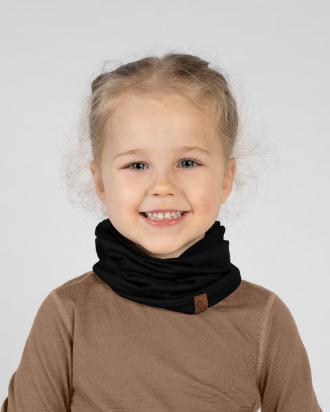 A studio portrait of a young blonde girl looking directly at the camera with a big, bright smile. She is wearing a solid black neck gaiter gathered comfortably around her neck, featuring a small brown leather logo patch on the lower edge. She is wearing a light brown long-sleeved shirt against a plain light grey background.