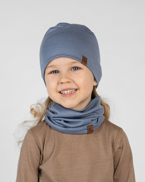 A close-up studio portrait of a smiling young blonde girl tilting her head slightly and looking directly at the camera against a plain light grey background. She is wearing a brown long-sleeved top and a matching set of a dusty blue beanie and a neck gaiter bunched around her neck. Both the beanie and gaiter have a small brown logo tag.