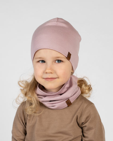 A close-up studio portrait of a young blonde girl looking off to her left against a plain light grey background. She is wearing a brown long-sleeved top and a matching set of a dusty pink beanie and a neck gaiter bunched around her neck. Both the beanie and gaiter have a small brown logo tag.