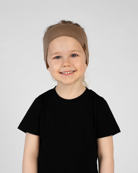 A close-up studio portrait of a smiling young girl looking directly at the camera against a white background. She is wearing a black short-sleeved t-shirt and a wide beige headband with a small brown logo tag on the side.
