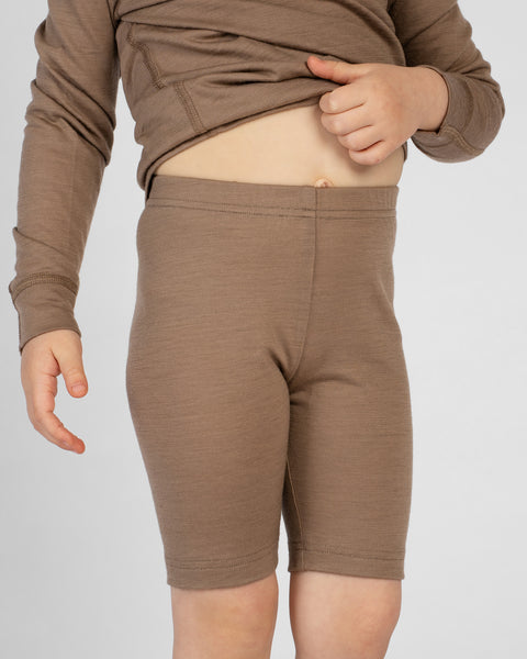 A close-up, slightly angled view of a child wearing form-fitting beige merino wool cycling shorts and a matching long-sleeve top against a plain light gray background. The child is gently lifting the hem of the top with one hand, exposing their midriff and the elastic waistband of the shorts.