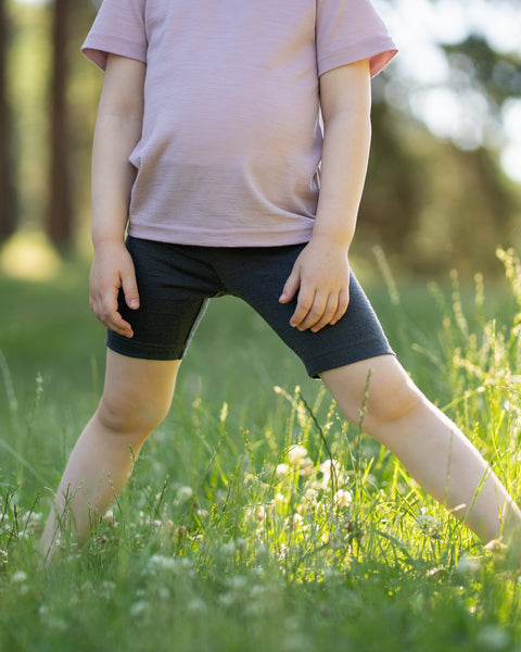 Close-up of a child walking through a grassy meadow wearing dark gray Merino wool biker shorts and a light pink T-shirt. The image highlights the soft, stretchy fit and breathable comfort of the kids' shorts for everyday outdoor adventures.