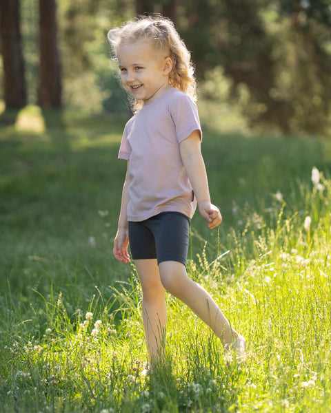 Close-up of a child standing outdoors in a grassy park wearing dark gray Merino wool biker shorts paired with a light pink Merino T-shirt.