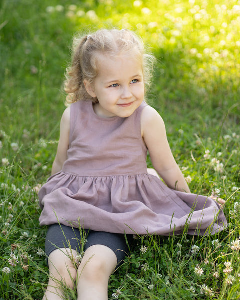 Young girl sitting on the grass in a sunny park wearing a dusty pink sleeveless dress with dark gray Merino wool biker shorts underneath. The image showcases the soft, breathable shorts as a comfortable layering piece for active outdoor play.