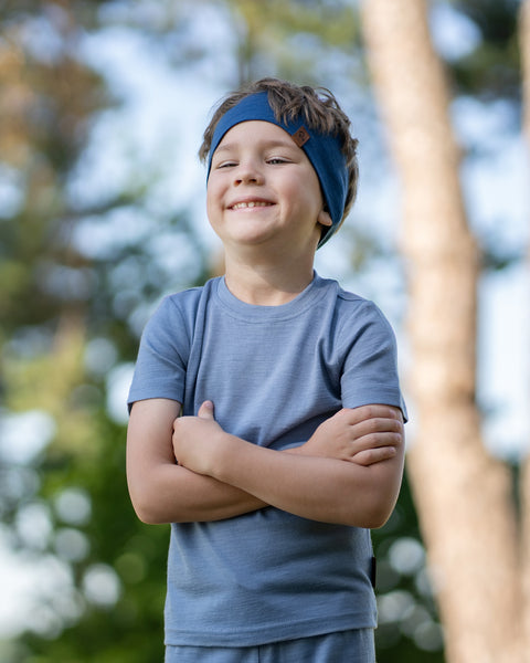 A young boy standing outdoors with his arms confidently crossed, smiling brightly. He is wearing a slate blue short-sleeve t-shirt and a dark blue headband, with blurred trees in the background.