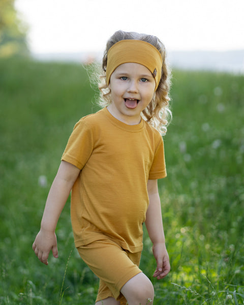 Young girl walking through a grassy meadow wearing a yellow Merino wool T-shirt, matching biker shorts, and a coordinating Merino wool headband. The soft, breathable outfit is showcased in a sunny outdoor setting, offering comfortable everyday wear for active kids.