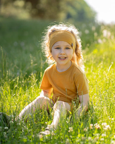 A smiling young girl with curly blonde hair sitting in a sunlit field of tall green grass and clover. She is wearing a matching mustard yellow short-sleeve t-shirt and a wide headband.