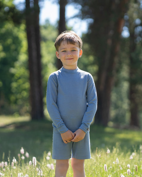 Young boy standing outdoors in a sunny park wearing a light blue long-sleeve Merino wool top and matching shorts. He poses confidently with his hands on his hips, showcasing the comfortable kids' Merino wool outfit against a softly blurred natural background.
