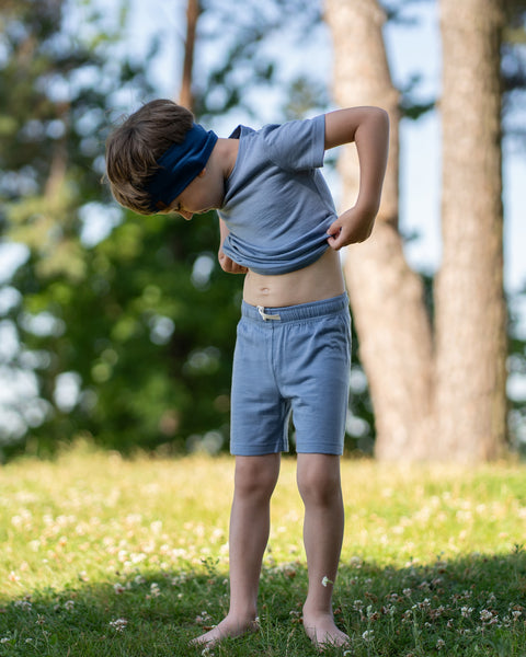 A young boy stands barefoot in a grassy field, looking down as he lifts the hem of his blue short-sleeve t-shirt, exposing his belly. He is wearing matching blue shorts with a white drawstring and a wide, dark blue headband. Trees are visible in the background.