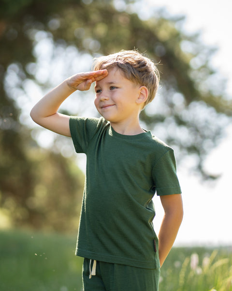 Young boy wearing a dark green Merino wool T-shirt and matching shorts. The soft, breathable kids' outfit is showcased in a sunny outdoor setting, perfect for active play and everyday comfort.