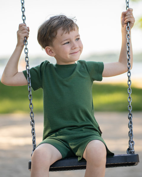 Young boy sitting on a playground swing wearing a forest green Merino wool T-shirt and matching shorts. The soft, breathable kids' outfit is showcased in a sunny outdoor setting, perfect for active play and everyday comfort.