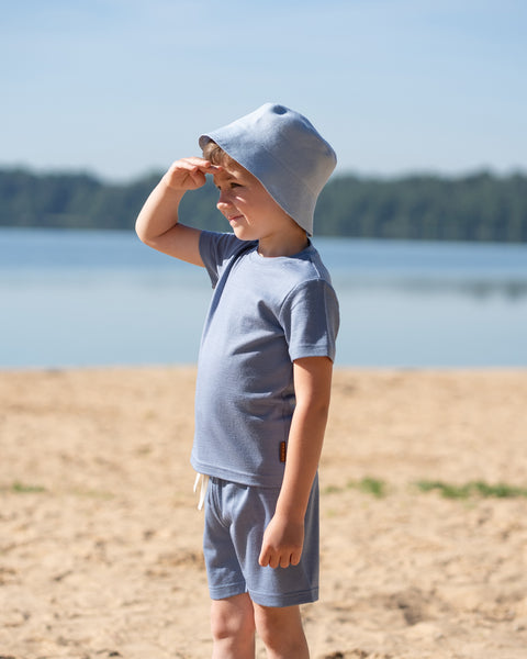 Young boy standing on a sandy beach wearing a dusty blue Merino wool T-shirt, matching shorts, and a bucket hat. The lightweight, breathable kids' outfit is showcased by the lakeside, making it ideal for sunny days, travel, and outdoor adventures.