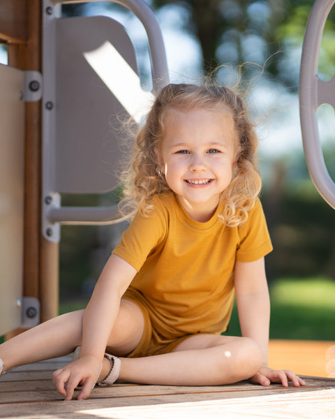 Young girl smiling on a playground wearing a yellow Merino wool T-shirt and matching shorts. The soft, breathable kids' outfit is showcased in a sunny outdoor setting, designed for comfortable movement during play and everyday adventures.