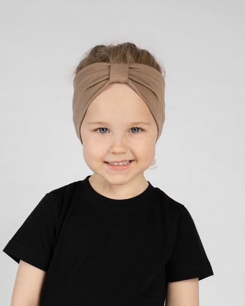 A close-up studio portrait of a young girl smiling directly at the camera against a plain light grey background. She is wearing a black short-sleeved t-shirt and a beige ribbon headband.