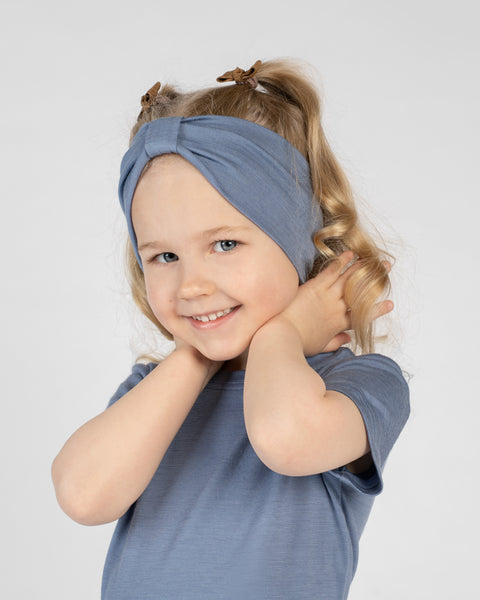 A close-up studio portrait of a young girl smiling directly at the camera. She wears a short-sleeved dusty blue top and a matching dusty blue ribbon headband. Her hands are gently resting behind her neck against a plain light grey background.