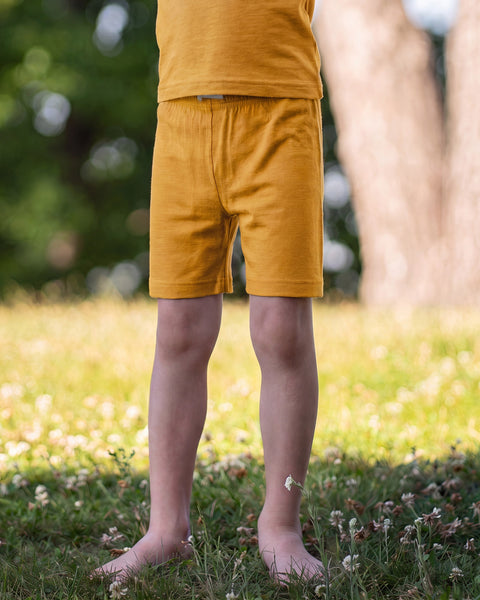 A close-up shot of a child's lower half standing barefoot in a grassy field with clover. The child is wearing simple mustard yellow shorts, with the hem of a matching yellow shirt visible at the top.