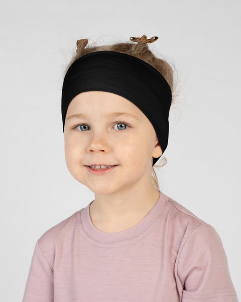 A close-up studio portrait of a smiling young child looking directly at the camera against a plain light grey background. She is wearing a light pink short-sleeved t-shirt and a wide black headband with the ribbon tie details visible at the top of her head.