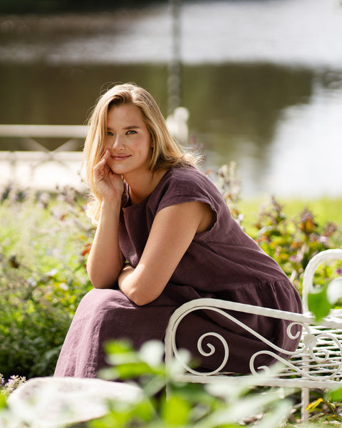 A woman sits comfortably on a decorative white bench by a lake, leaning forward with her arms crossed over her knees. She wears a shadow purple short-sleeve linen dress and smiles softly directly at the camera.