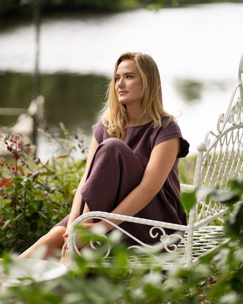 A barefoot woman sits on an ornate white metal bench in a lush garden overlooking a lake. She gazes thoughtfully into the distance while wearing a shadow purple short-sleeve linen dress.