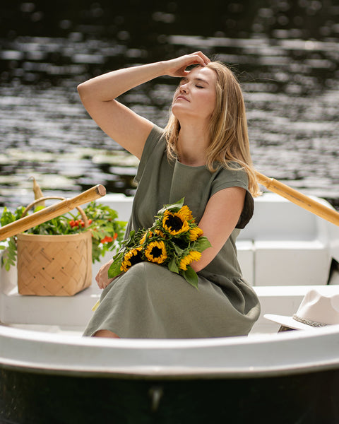 A woman wearing a relaxed stone green linen dress sits in a rowboat, holding a bright bunch of sunflowers. She has her eyes closed and her face turned upward toward the sunlight, resting one hand softly on her forehead in a peaceful pose.