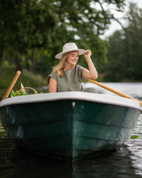 A smiling woman wearing a stone green linen dress and a light-colored hat sits comfortably in a small rowboat. She rests one arm gracefully on the side of the boat as it gently drifts on the calm water, surrounded by lush green trees on the far bank.