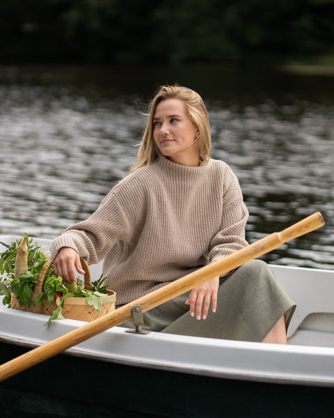 A woman relaxes in a small rowboat, wearing a beige mock-neck cashmere sweater over a stone green skirt. She is reaching into a woven basket full of fresh greens, with one hand casually resting on the wooden oar as she gazes thoughtfully over the water.
