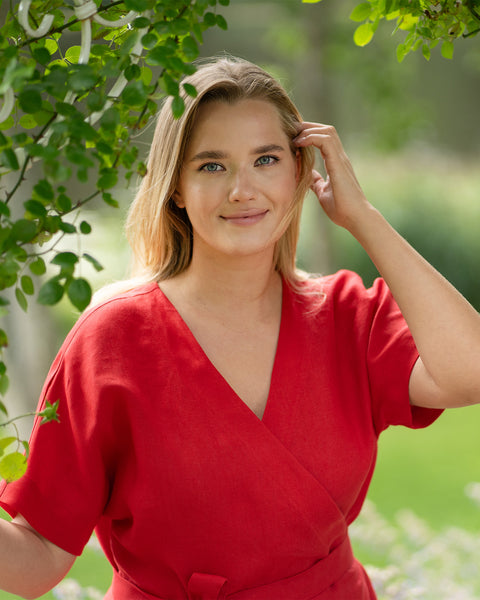 A blonde woman stands outdoors framed by green leaves, smiling warmly at the camera with one hand gently touching her hair. She is wearing a bright red short-sleeve linen wrap dress with a v-neckline and tie waist.